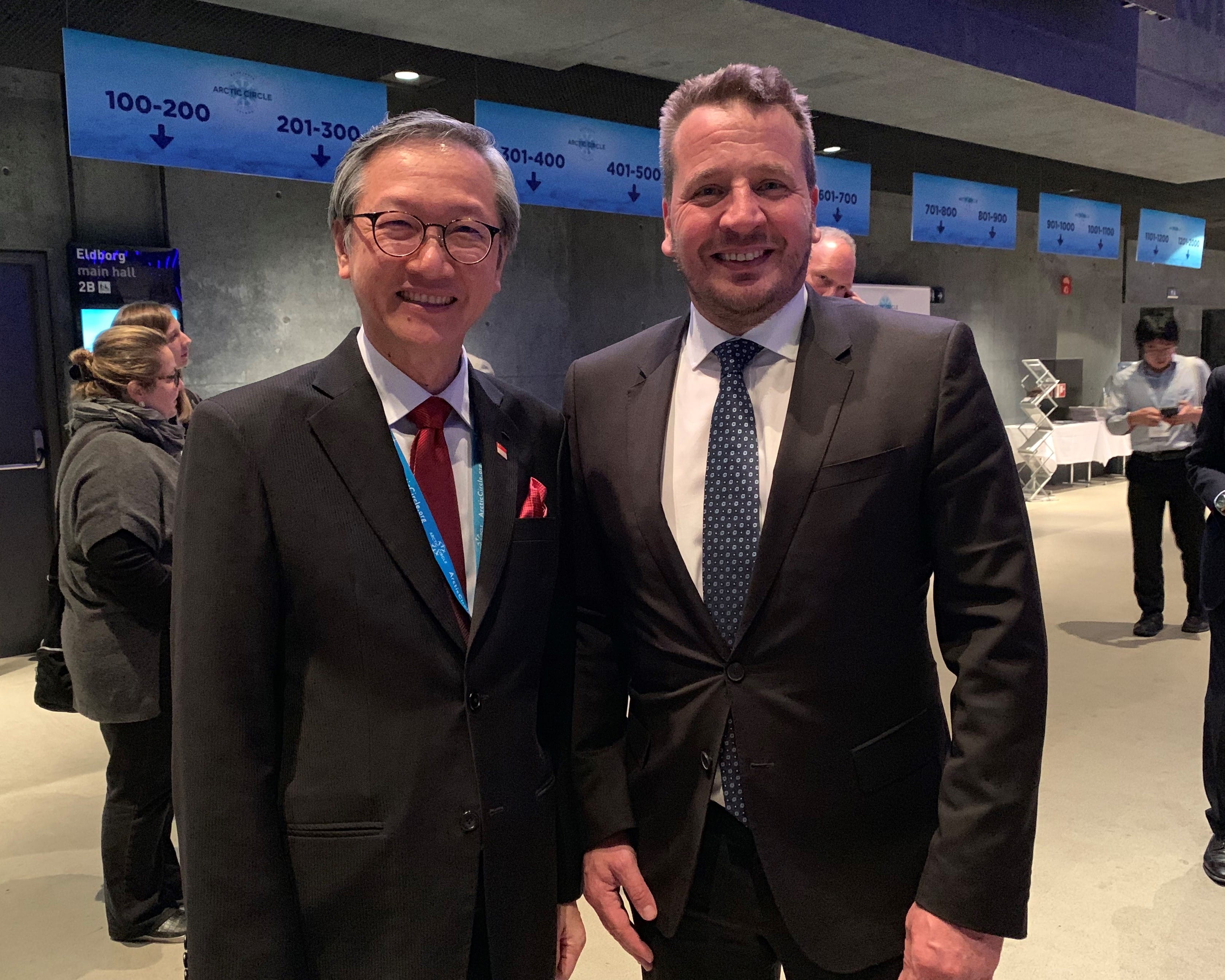 Two men in suits standing at an Arctic Circle event with hall directional signs.